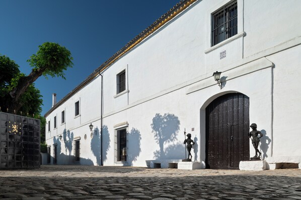 A cobblestone street with a white building featuring a large black door and a statue of a figure holding a staff. - Hospedium Hotel Cortijo Torre De La Reina (Guillena)