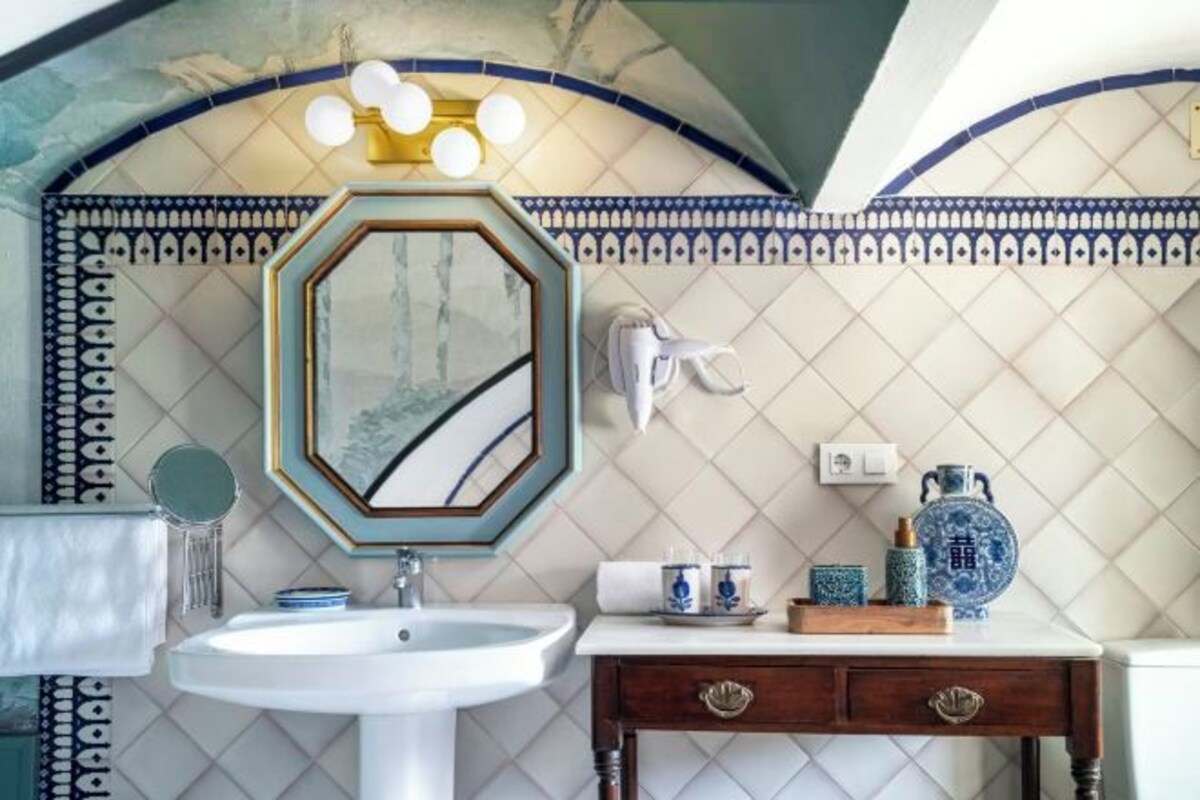A bathroom with a white sink, a mirror with a decorative frame, a vanity with a wooden cabinet, and a wall with patterned tiles.