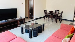 A living room with a flat-screen TV, a glass-top coffee table, a dining table with chairs, and a red sofa. - Muteithia Mbaabu (Nairobi)