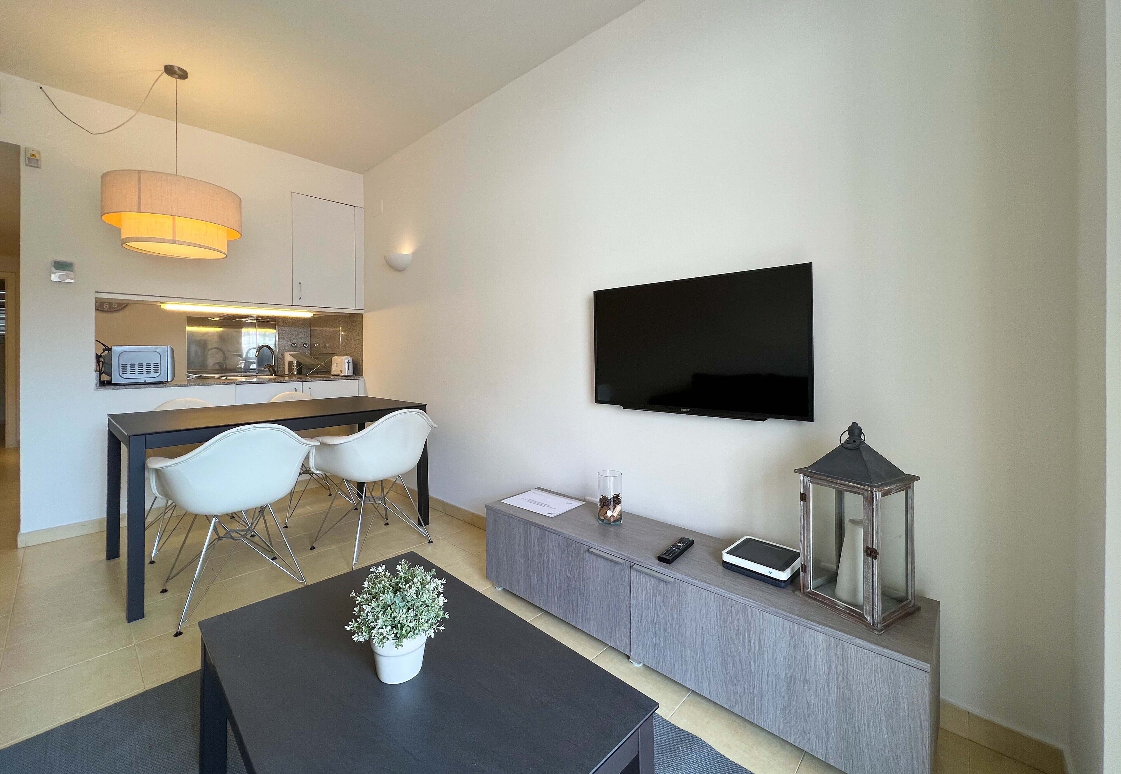 A modern living room with a flat-screen TV, a dining area with a kitchen in the background, and a grey console table with a lantern.