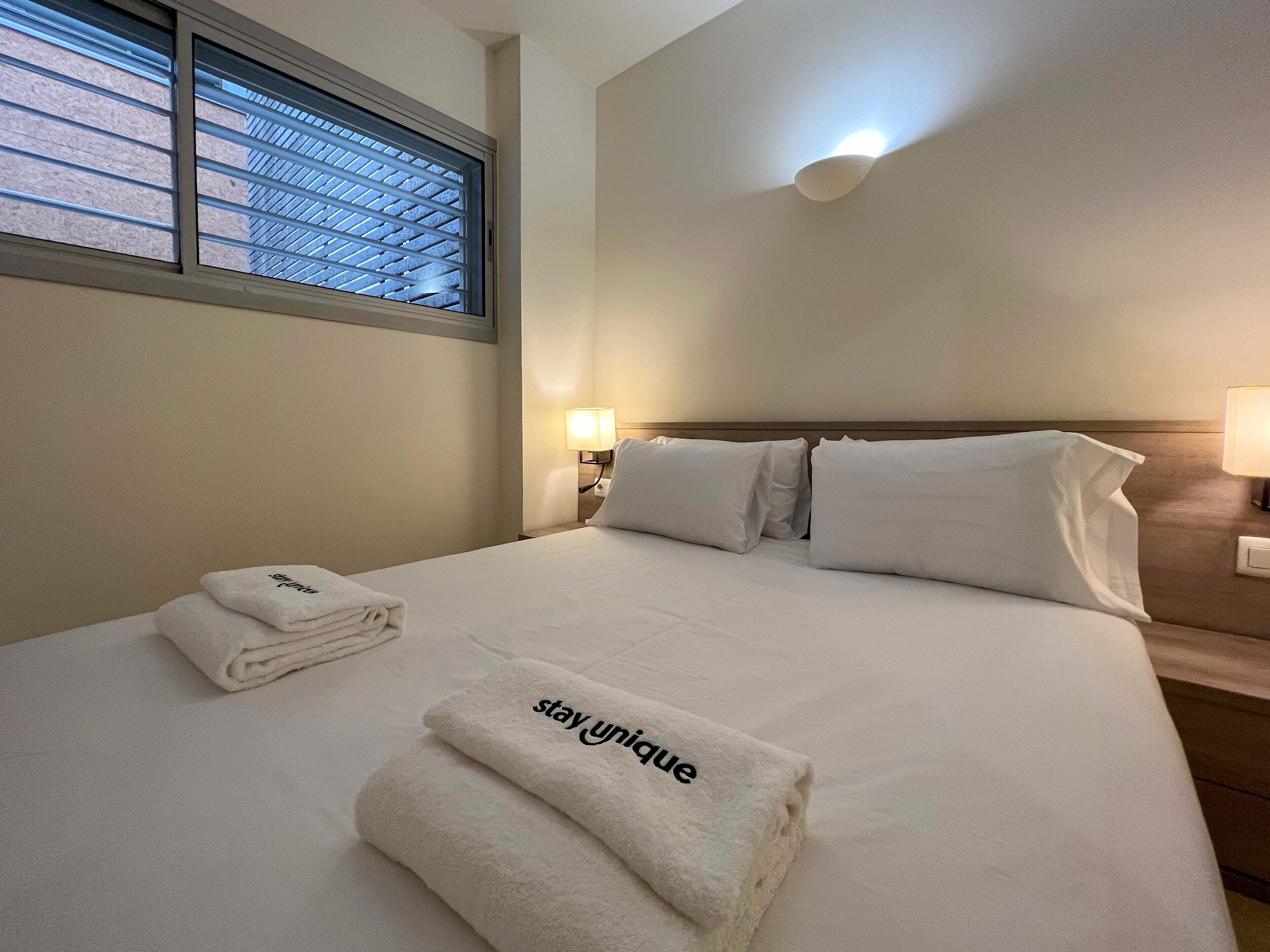 Una habitación de hotel con una cama bien hecha, una ventana con persianas, una lámpara fijada a la pared y una toalla doblada con el texto 'stay unique'.