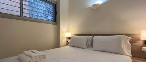 A hotel room with a neatly made bed, a window with blinds, a wall-mounted lamp, and a folded towel with "stay unique" text.