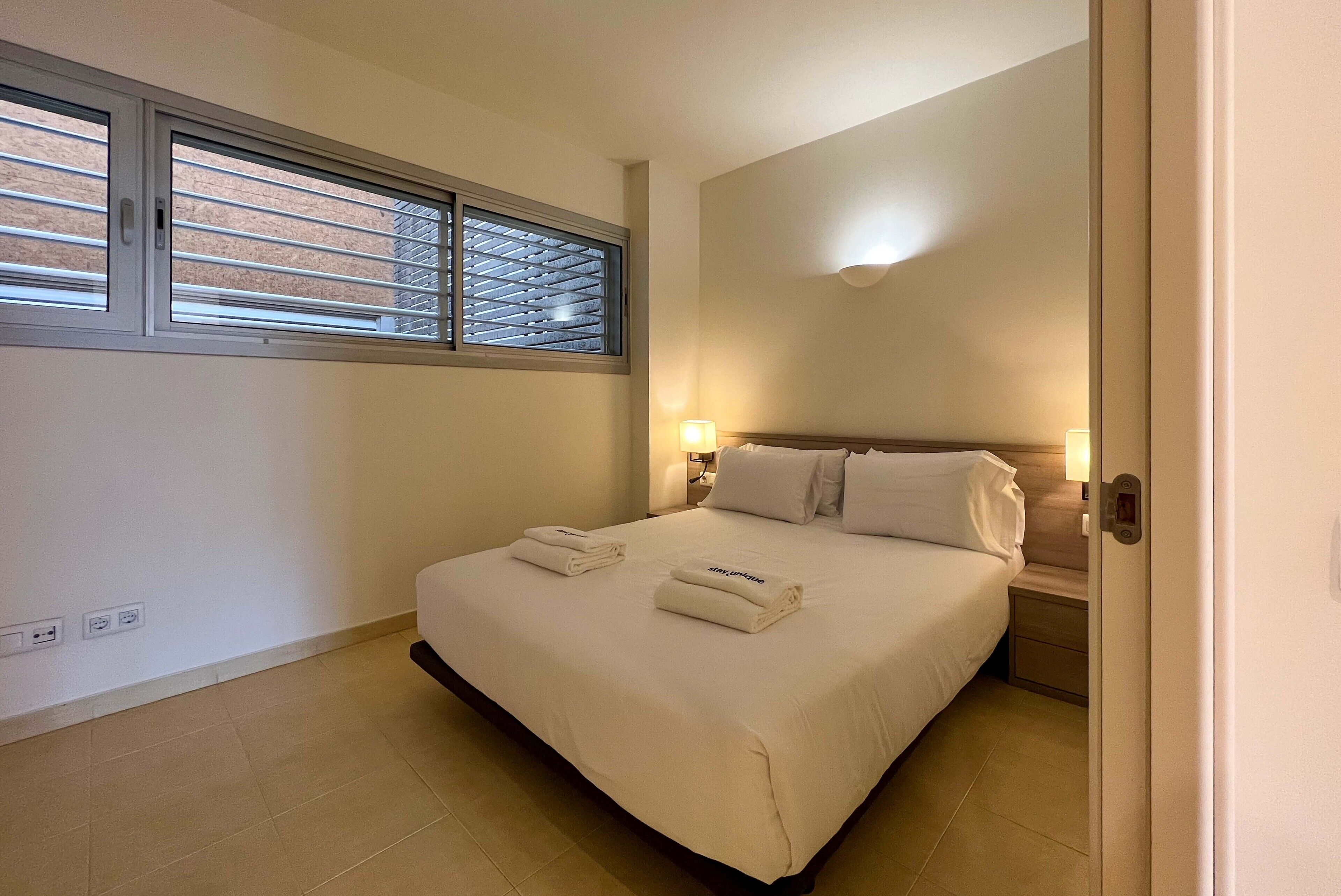 Une chambre d’hôtel avec un lit, deux lampes de chevet, une fenêtre avec des stores et une porte.