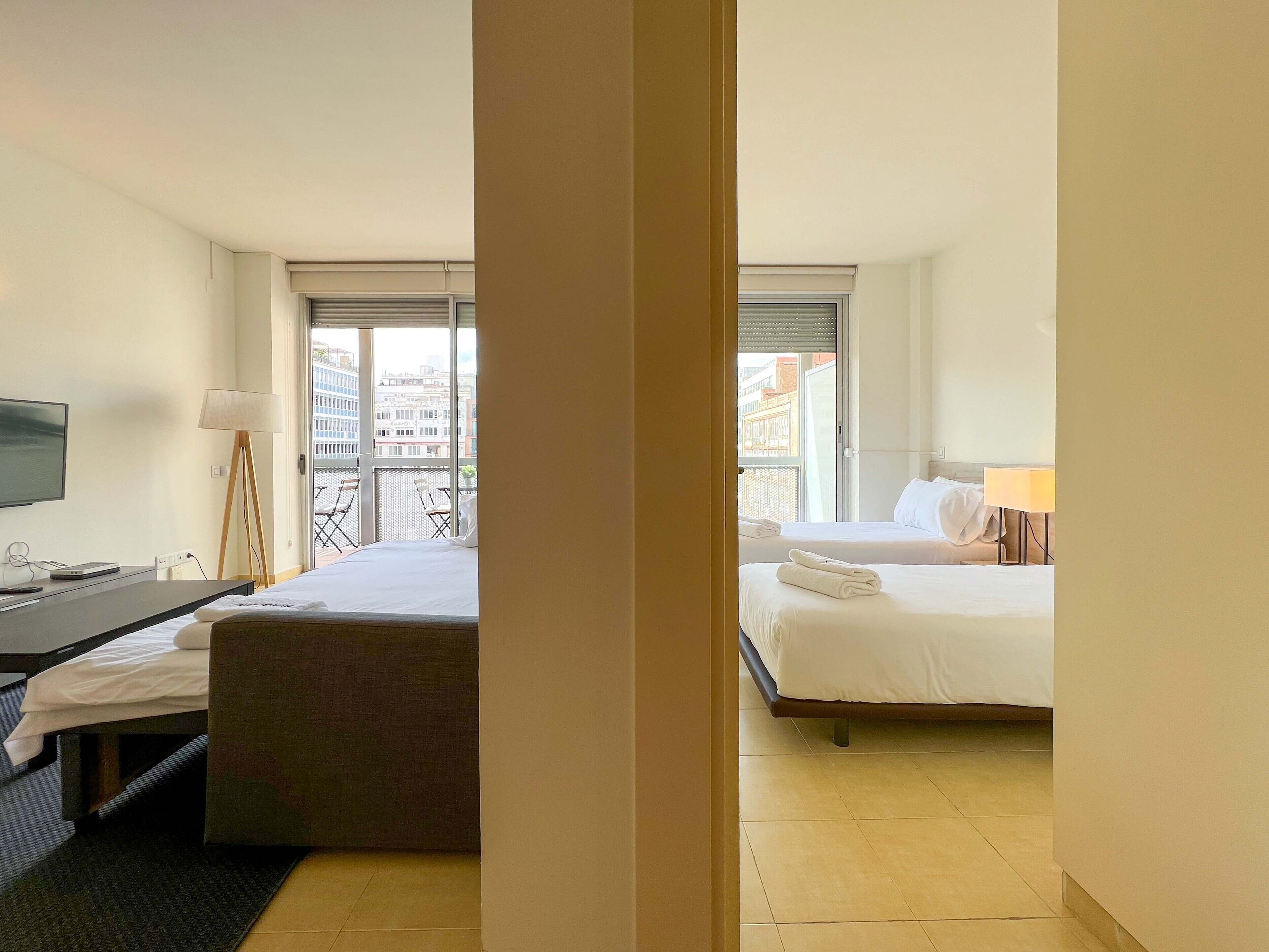 A hotel room with a bed, a desk, a chair, a TV, and a balcony with a city view.