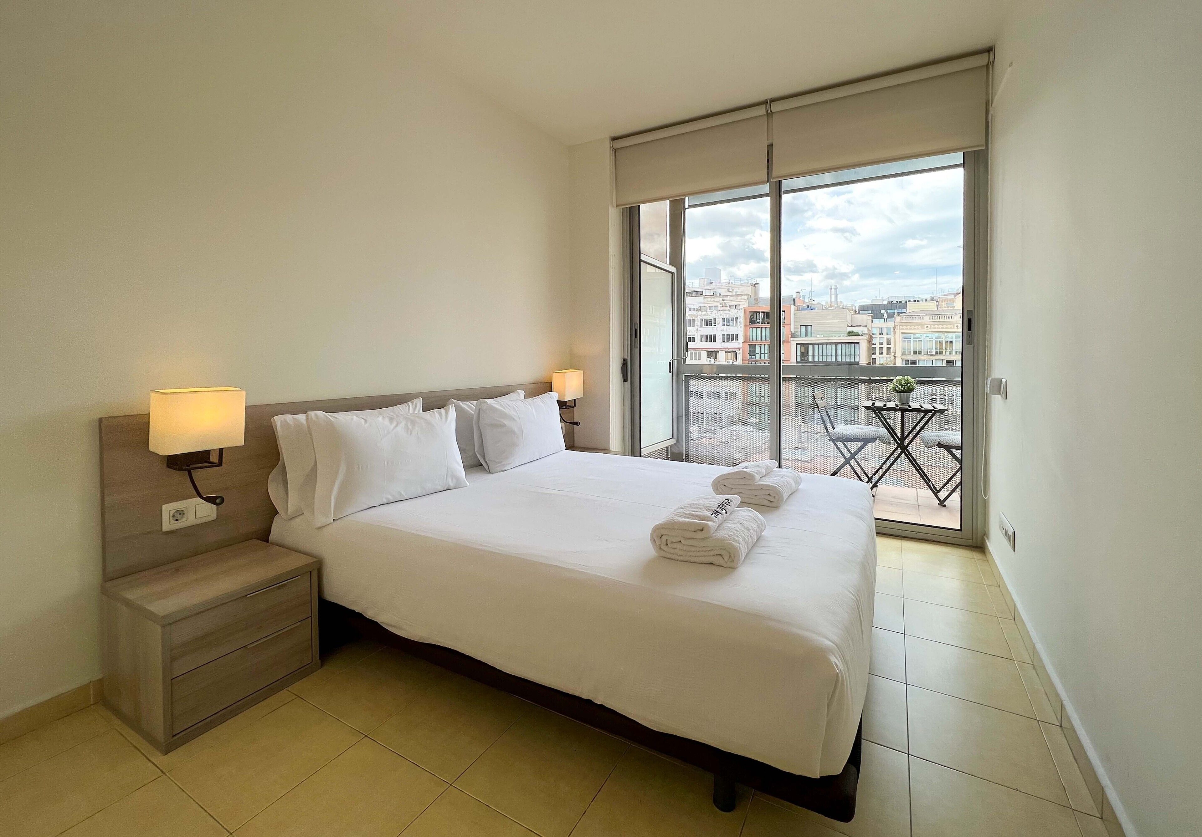 Una habitación de hotel con una cama grande, dos lámparas de noche, una mesita de noche y un balcón con vistas a la ciudad.
