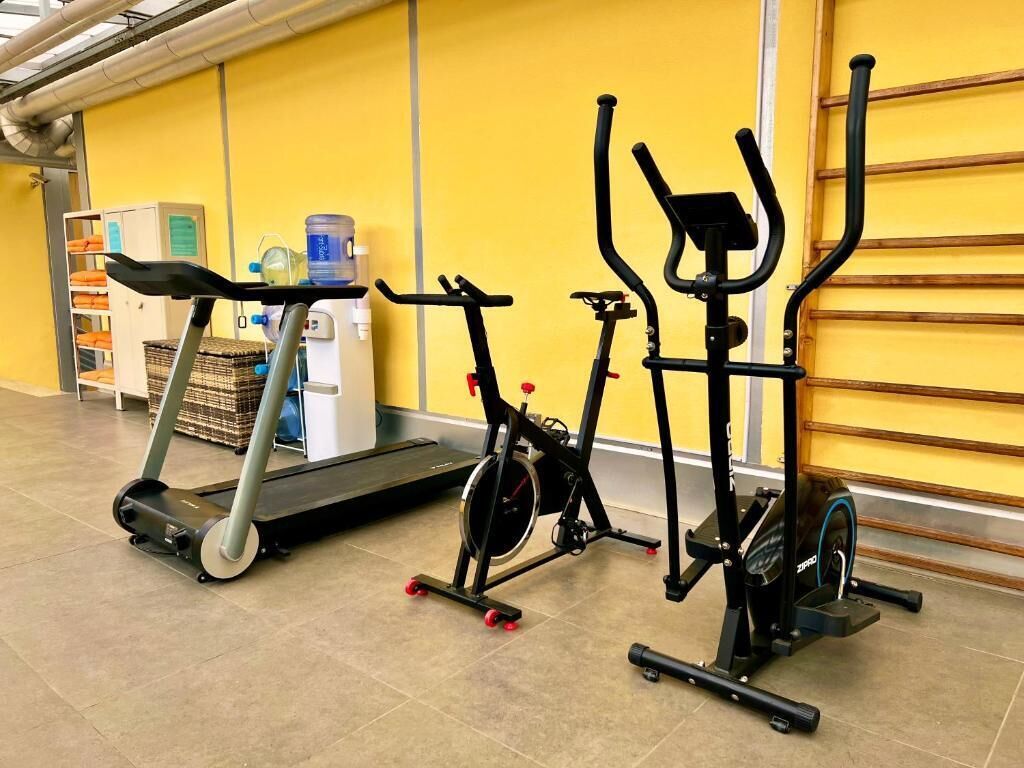 Three stationary bikes in a gym with a yellow wall and a treadmill in the background.