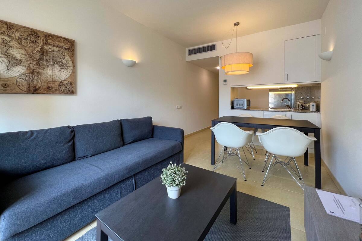 Ein modernes Wohnzimmer mit einem dunkelgrauen Sofa, einem Couchtisch mit Pflanze sowie einem Essbereich mit weißen Stühlen und einer Küche im Hintergrund.