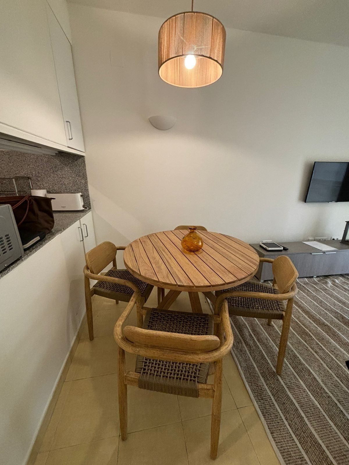 A compact living space with a round wooden dining table surrounded by four wicker chairs, a modern pendant light, a flat-screen TV mounted on the wall, and a small kitchen area with a microwave and a toaster.