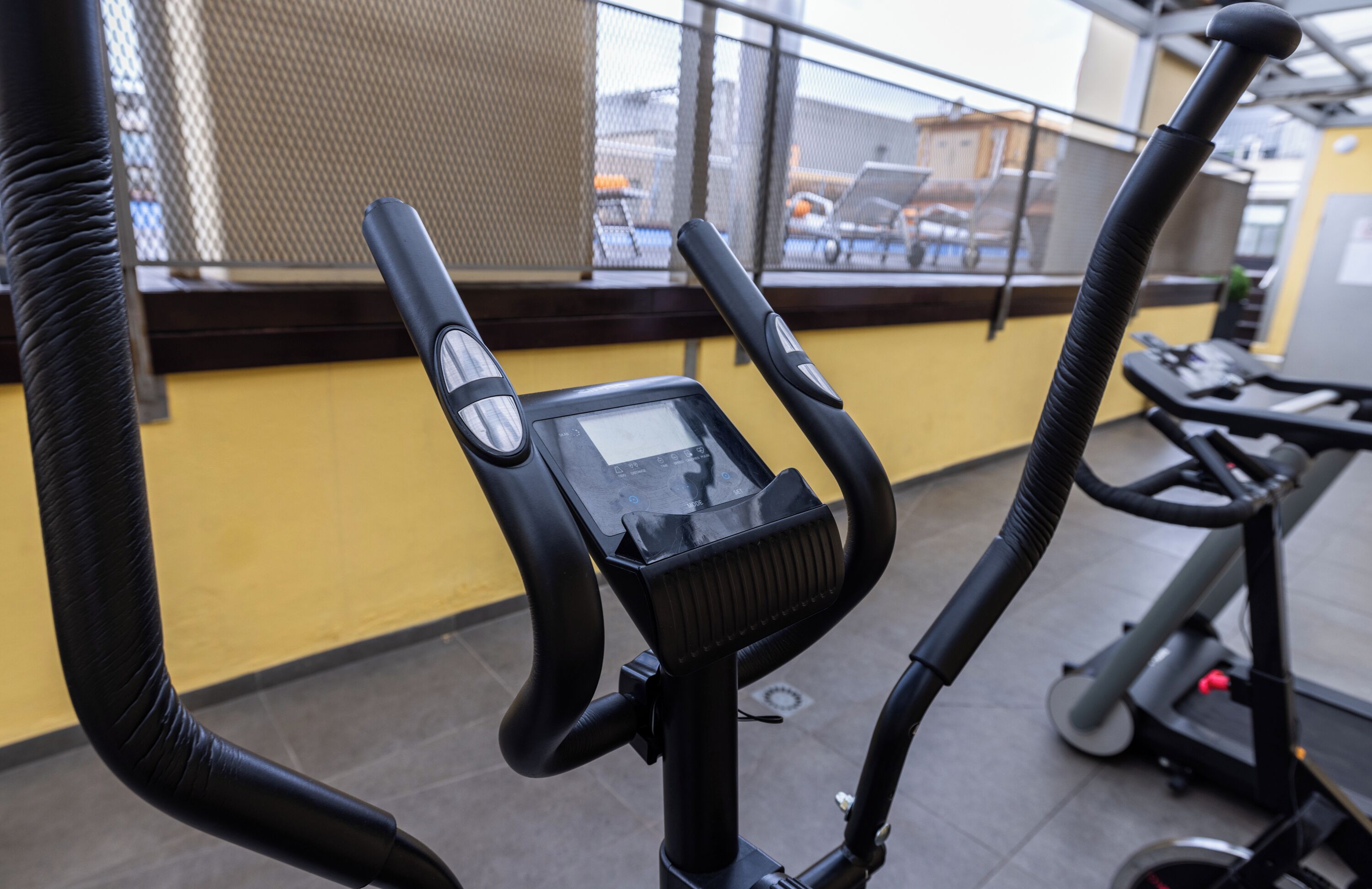 Un vélo d’appartement doté d’un écran numérique, un mur jaune et une vue sur les voitures garées à travers une fenêtre.