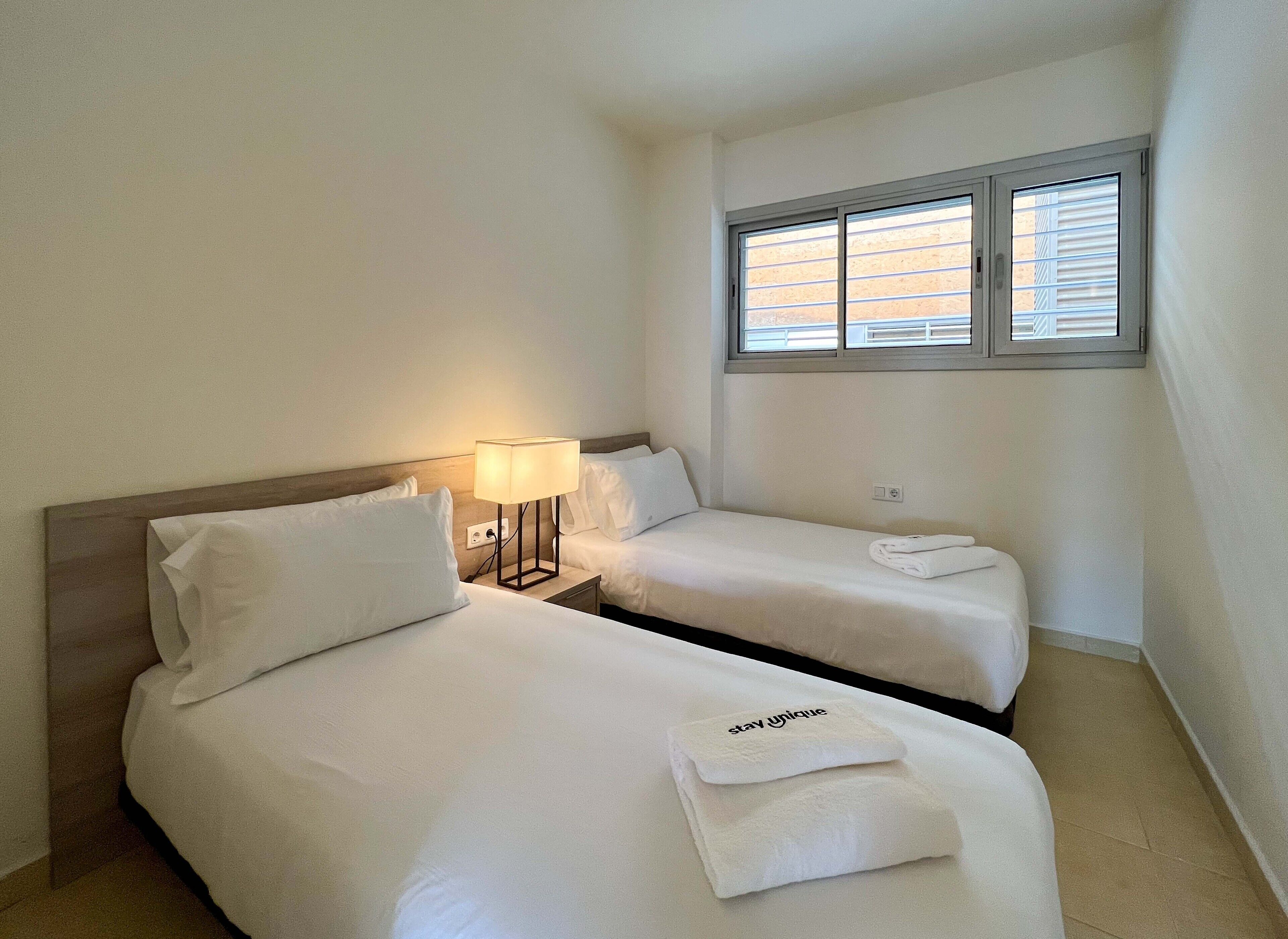 Ein Hotelzimmer mit zwei Einzelbetten, einem Nachttisch mit Lampe, einem Fenster mit Jalousien und einem Handtuch auf einem Bett.