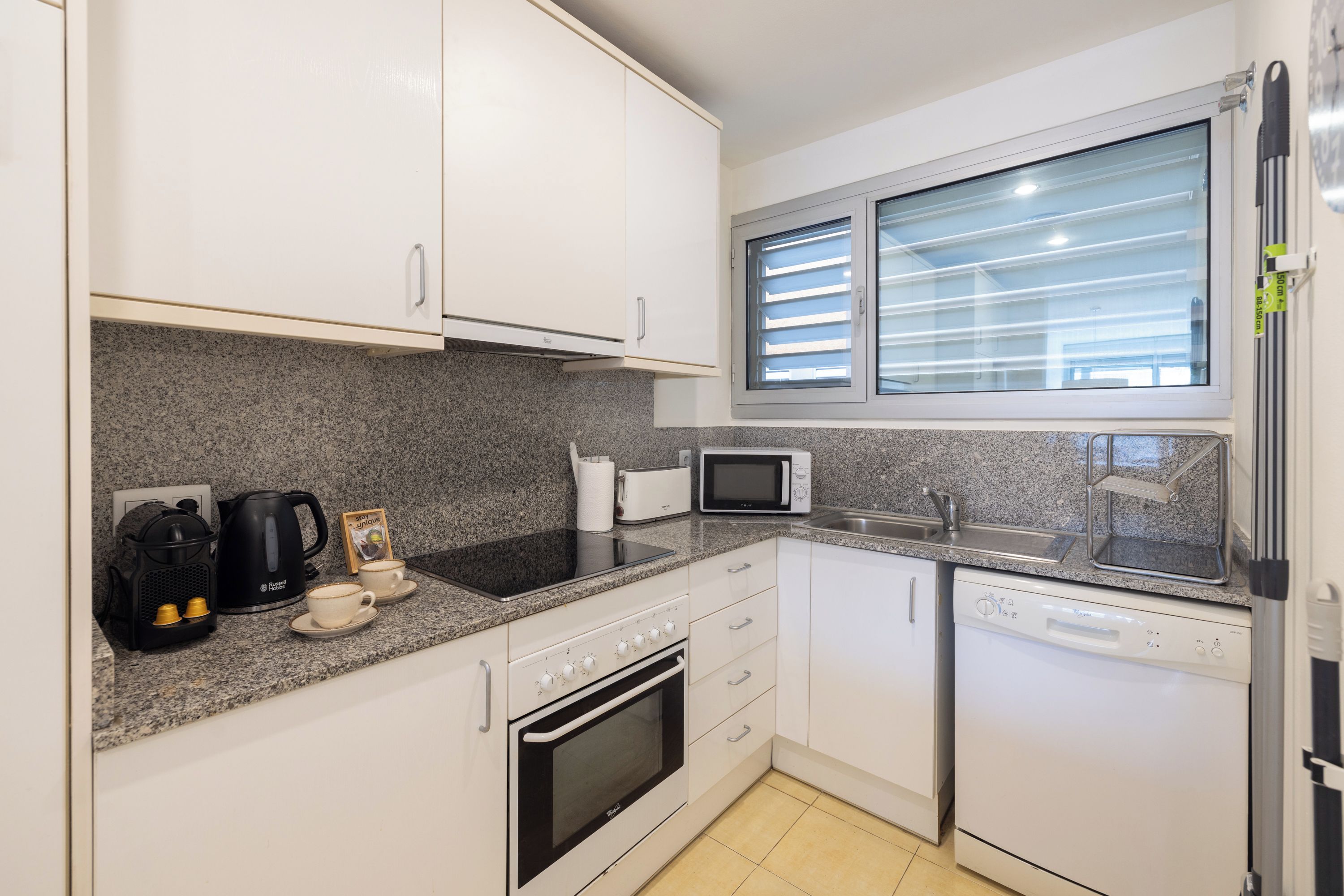 A compact kitchen with white cabinets, a granite countertop, a microwave, a toaster, a kettle, and a dishwasher.
