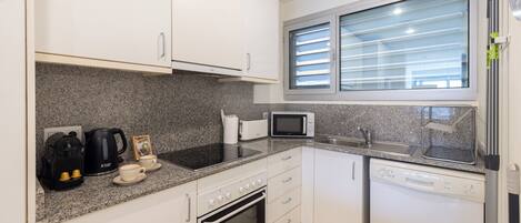 A compact kitchen with white cabinets, a granite countertop, a microwave, a toaster, a kettle, and a dishwasher.