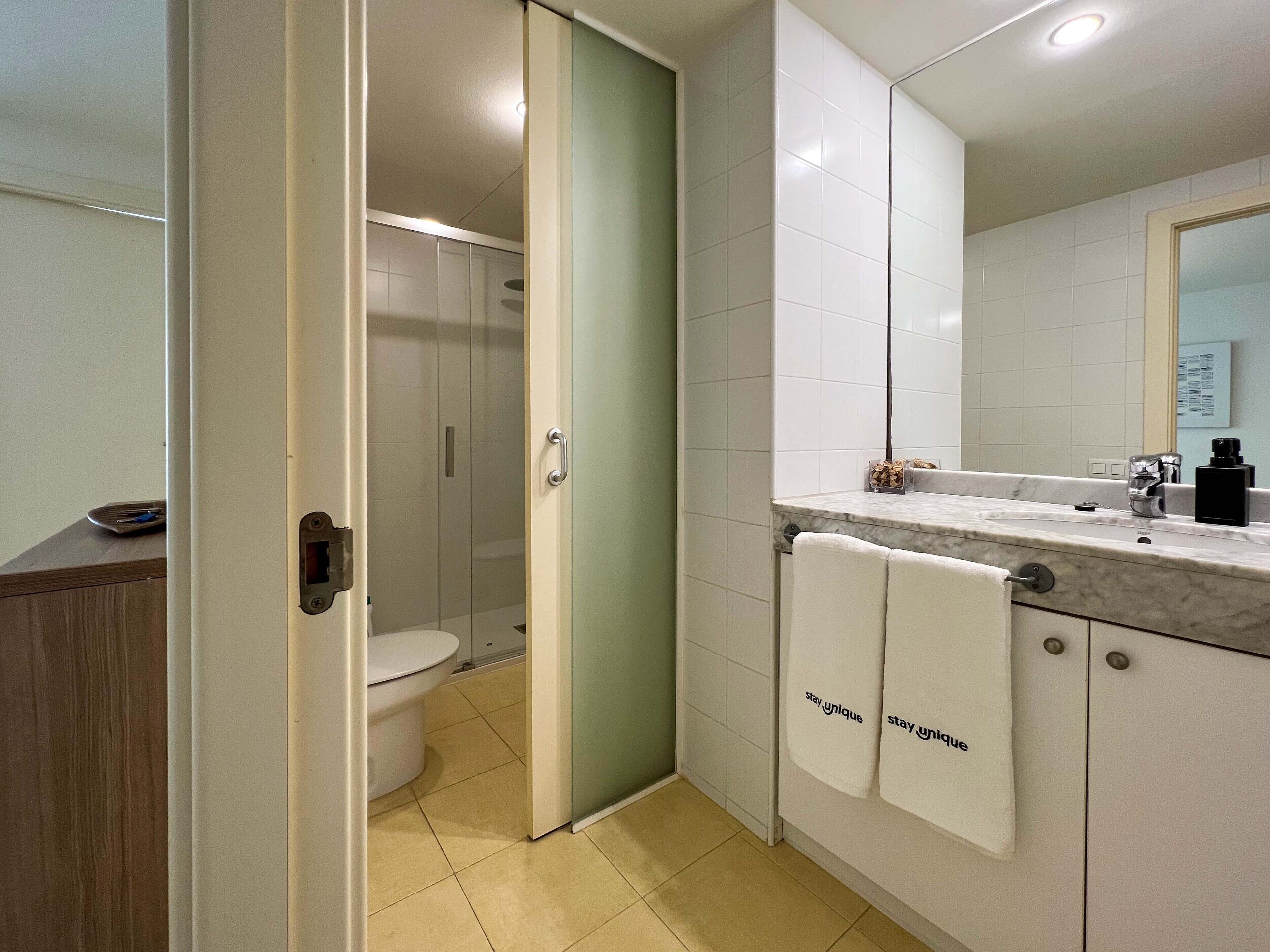 Ein modernes Badezimmer mit Duschkabine aus Glas, Toilette und Marmorwaschbecken. Zwei Handtücher hängen an der Wand.