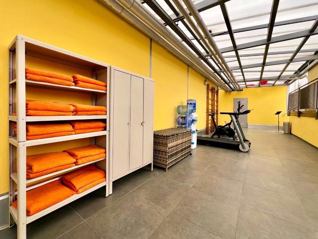 A room with a yellow wall, a white shelf unit with folded towels, a water dispenser, a trash bin, and a treadmill.