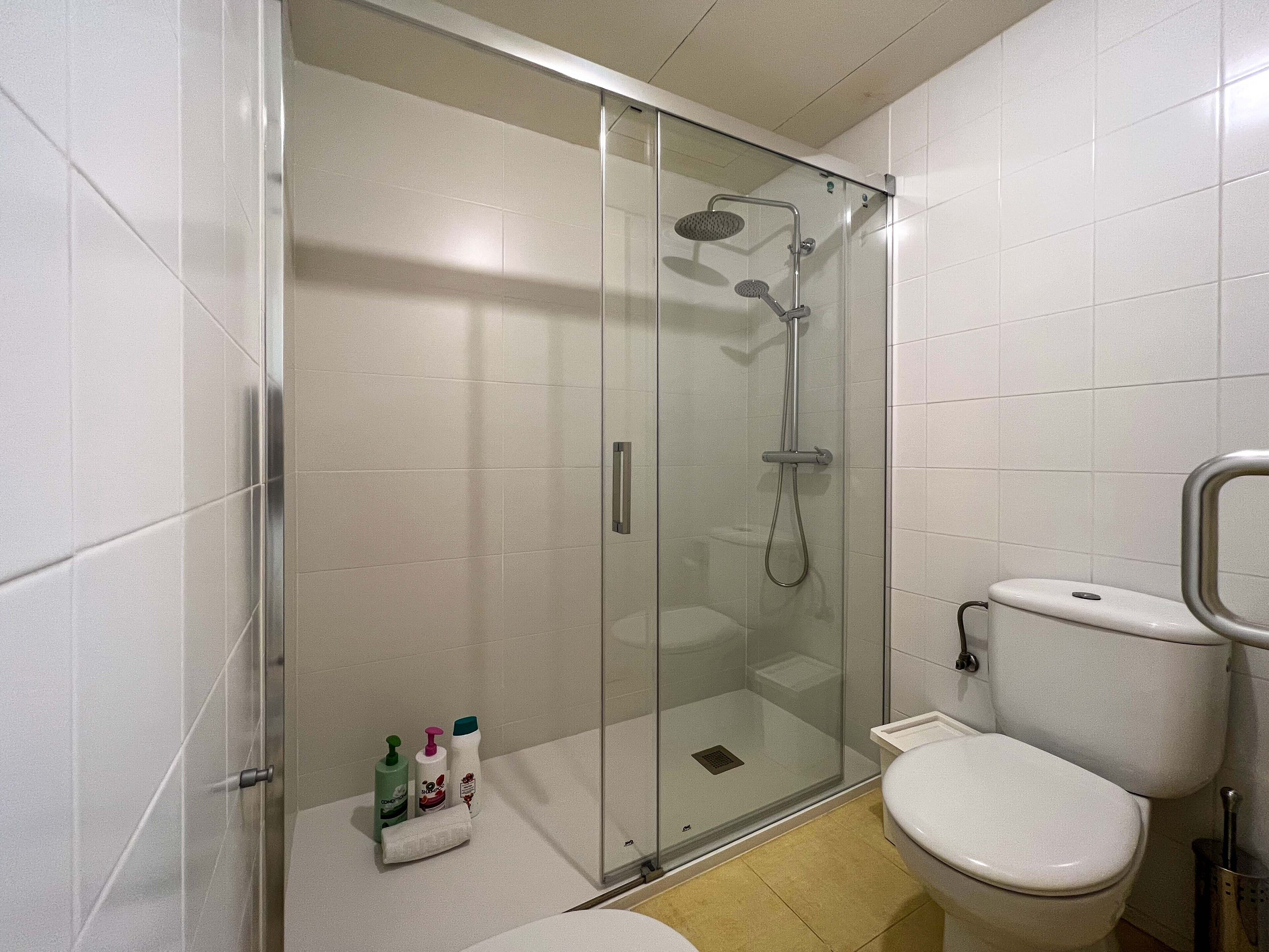 Un baño moderno con ducha envuelta en vidrio, inodoro y un estante pequeño con artículos de aseo.