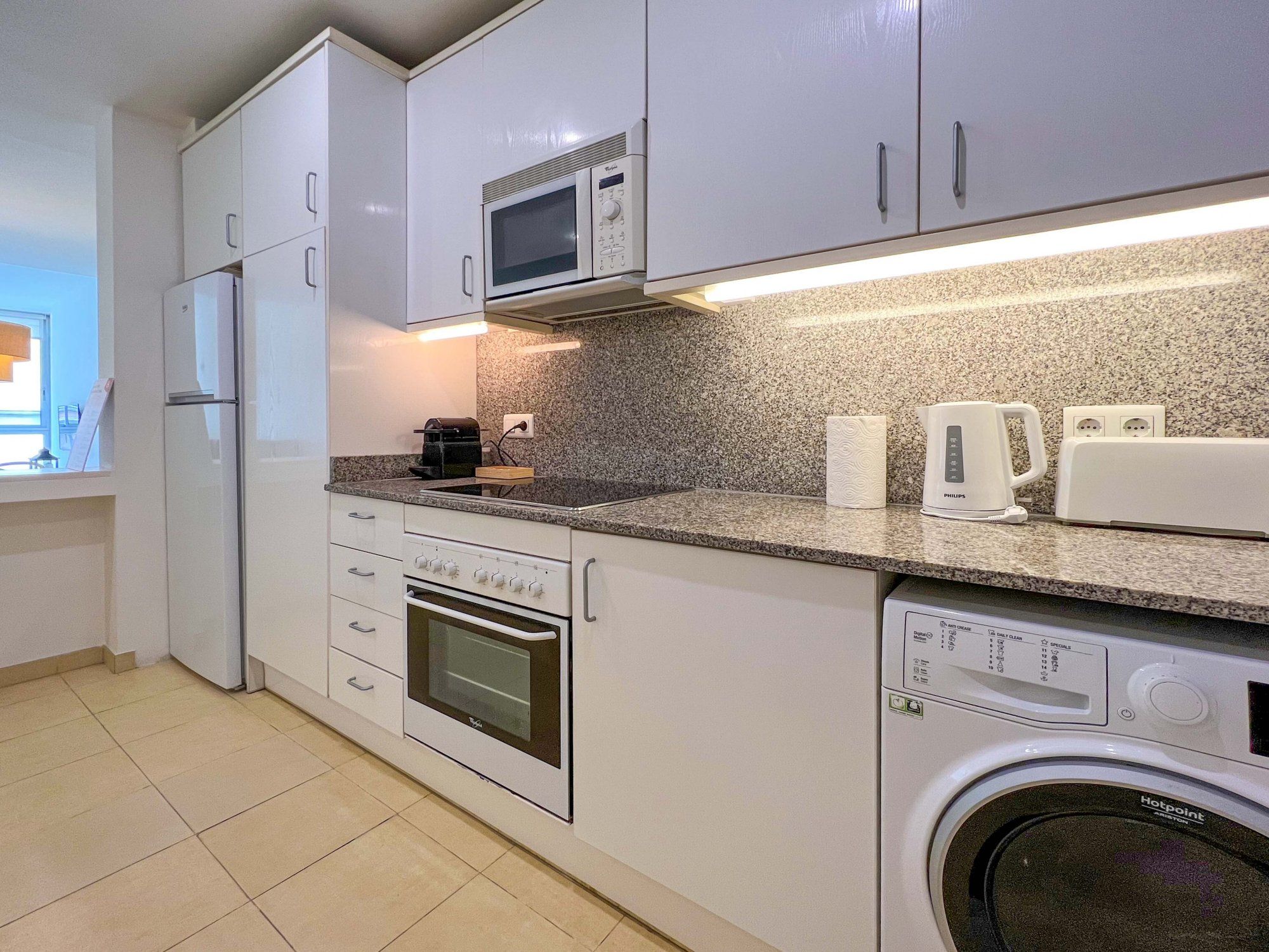 Eine kleine Küche mit weißen Schränken, einer Mikrowelle, einem Toaster, einer Kaffeemaschine, einem Backofen und einer Waschmaschine.