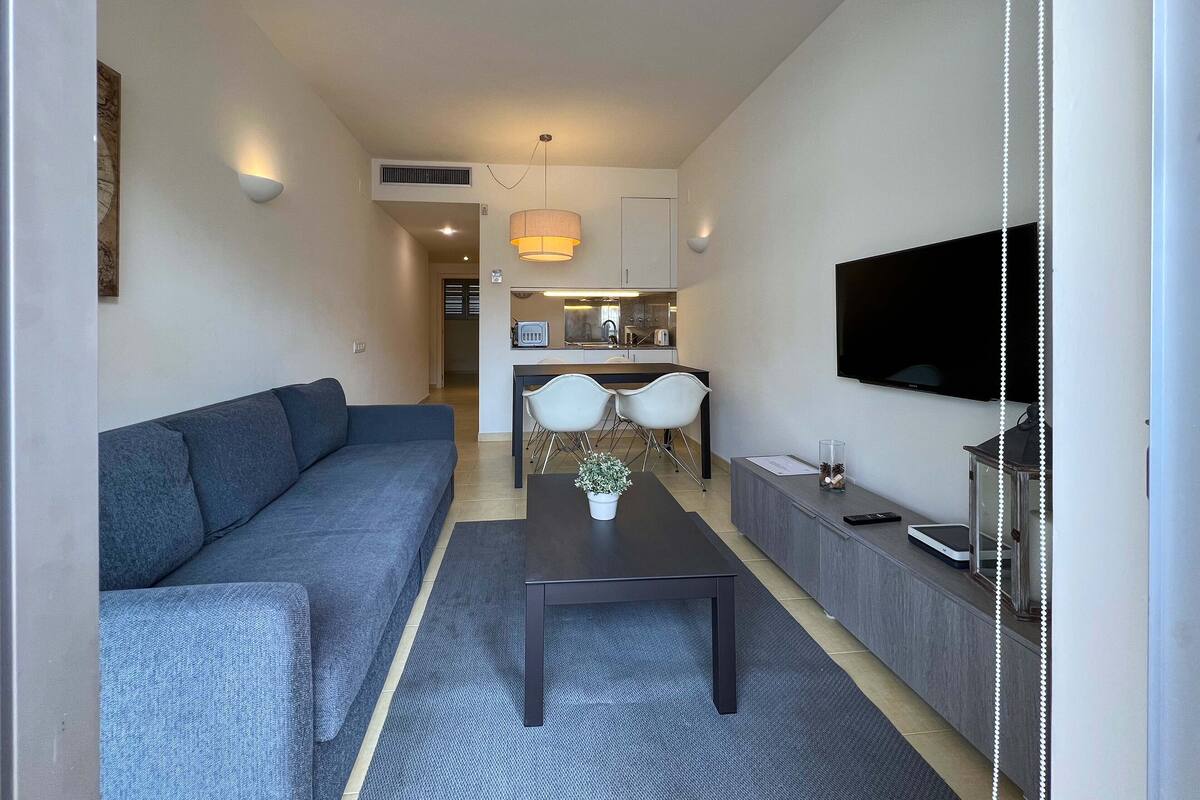 Ein modernes Wohnzimmer mit einer Couch, einem Couchtisch und einem Fernseher. Im Hintergrund ist eine Küchenzeile mit einem Esstisch und Stühlen zu sehen.