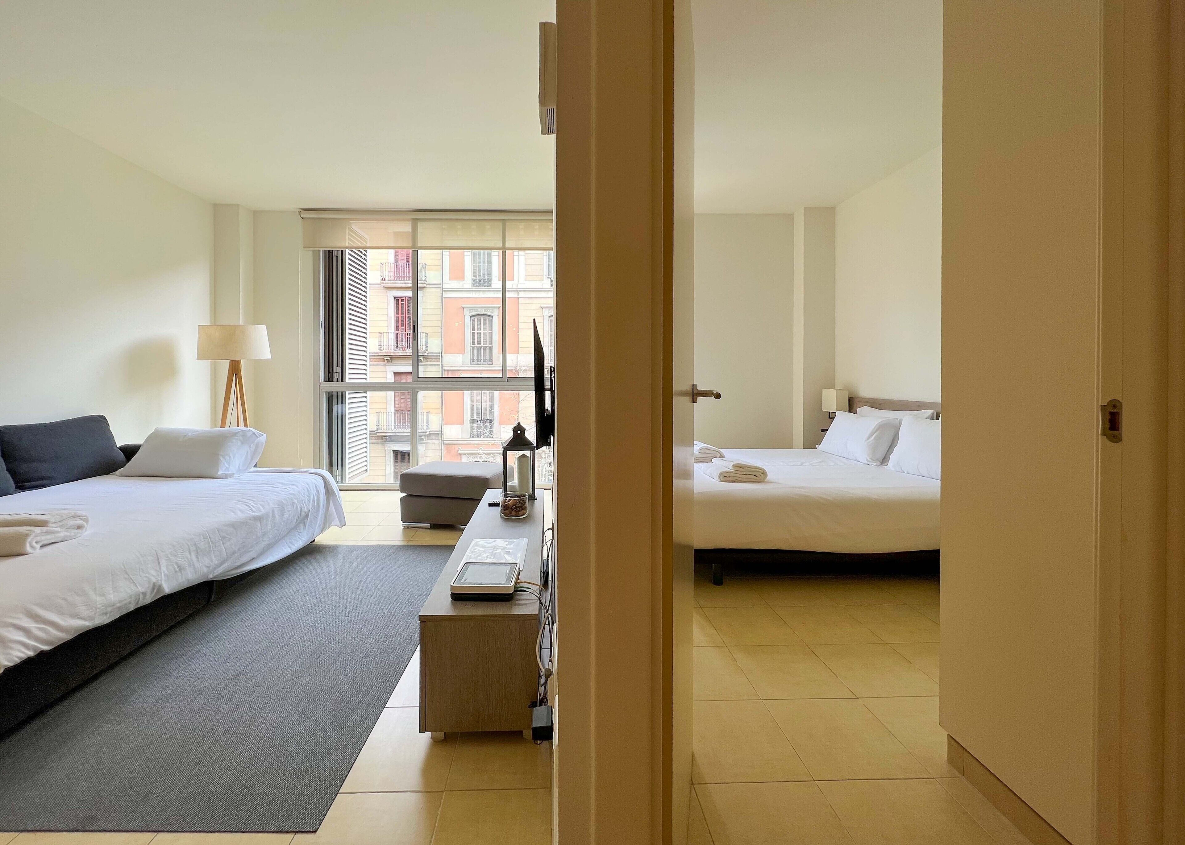 Ein Hotelzimmer mit zwei Betten, einem Schreibtisch, einer Lampe und Blick auf die Stadt durch große Fenster.
