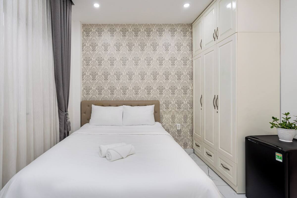A hotel room with a bed, curtains, a wardrobe, and a small refrigerator.