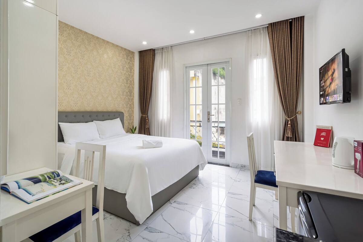 A hotel room with a bed, a desk, a chair, a TV, and a balcony with curtains.