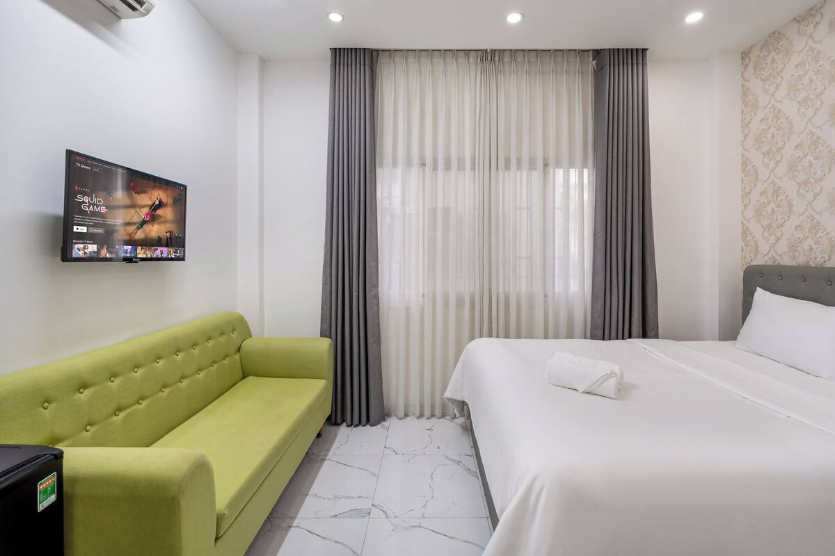A hotel room with a bed, a green sofa, a TV mounted on the wall, and a window with curtains.