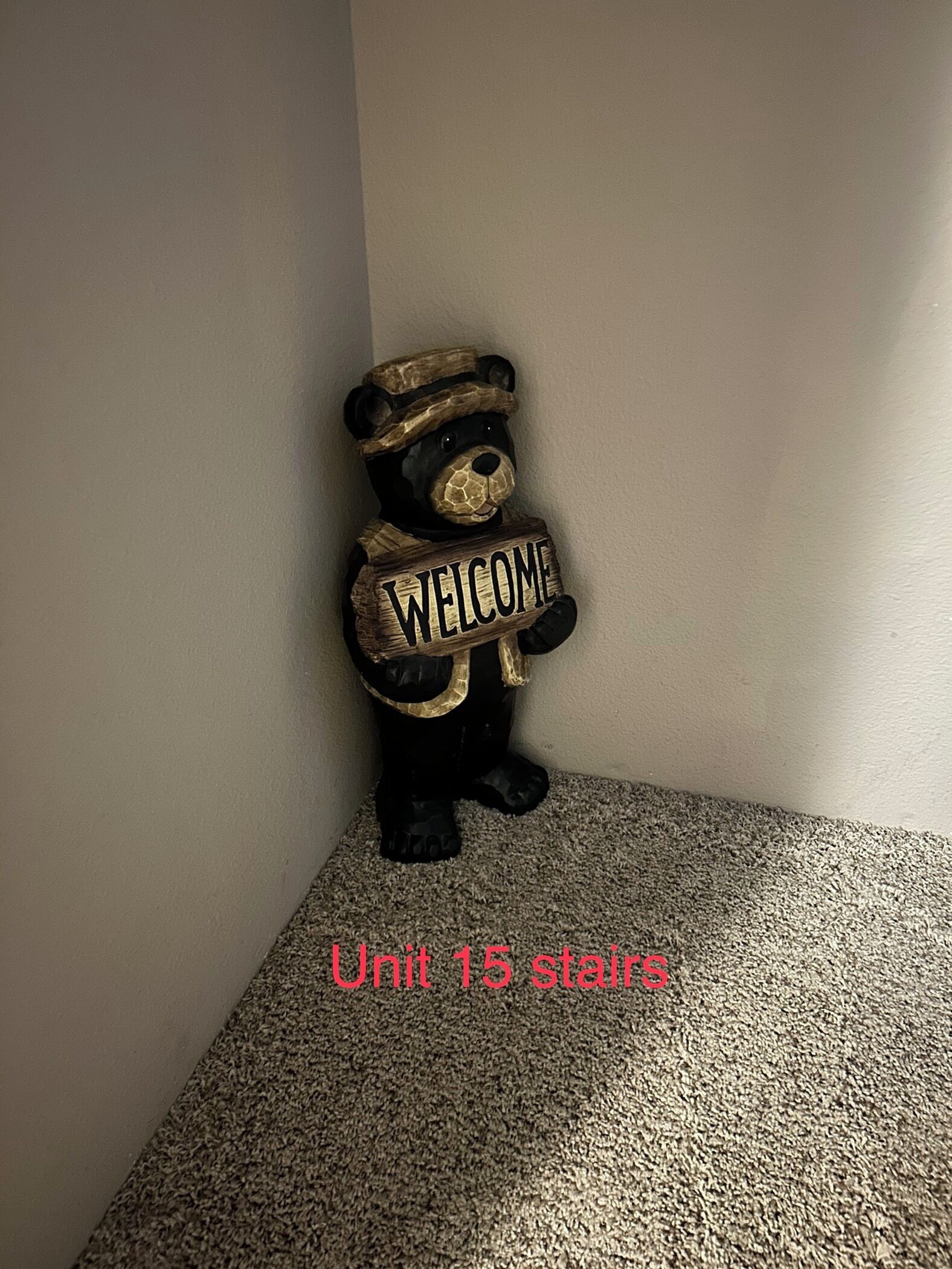 Una decoración con forma de oso que incluye un cartel con la palabra 'WELCOME', ubicada en una esquina con piso alfombrado.