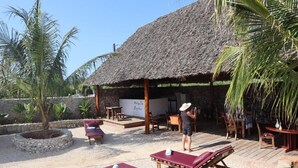 Un’area esterna con tetto di paglia, poltrone relax e un palco, circondata da palme e un muro in pietra.