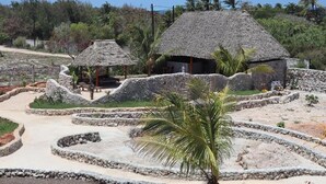Una casa tradizionale con tetto di paglia, con un vialetto in pietra che vi conduce, circondata da palme e un paesaggio collinare.