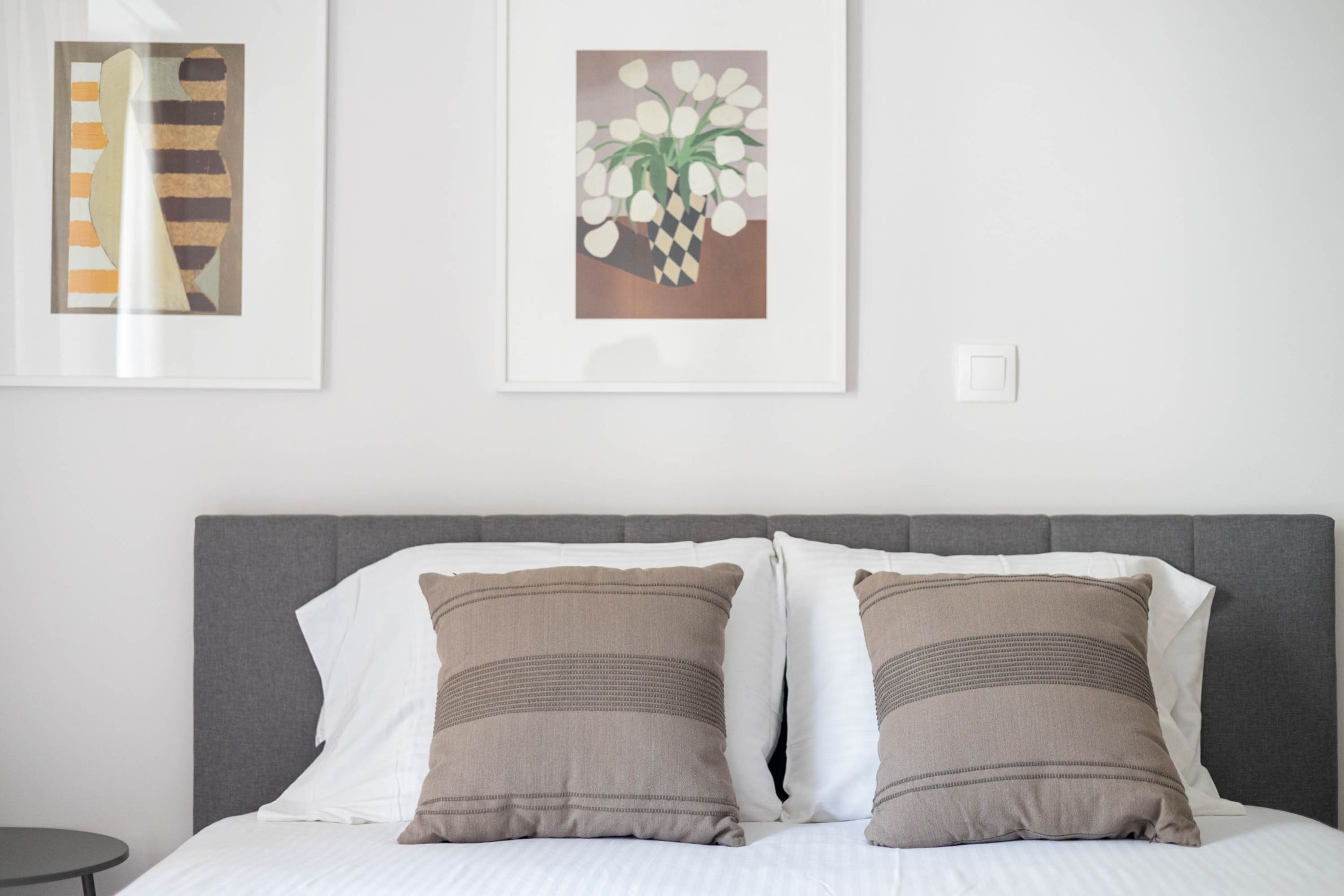 Una cama bien hecha con dos almohadas, un cabecero gris y dos cuadros enmarcados en la pared.