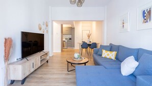 A modern living room with a flat-screen TV, a blue sofa, a coffee table, and a dining area in the background. - Urban Comfort 2 Bdrm Next to Metro (Athens)