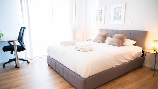 A neatly made bed with pillows and a bedside table with a lamp, a chair, and a desk with a plant. - Urban Comfort 2 Bdrm Next to Metro (Athens)
