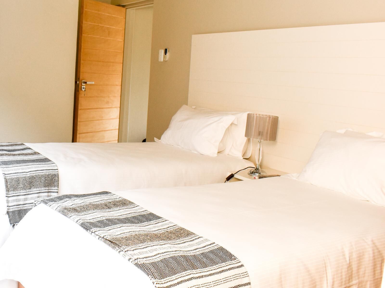 A hotel room with two beds, a wooden door, a bedside table with a lamp, and a wall-mounted thermostat.