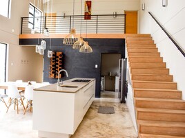 Una cocina moderna con una isla central, un comedor con sillas blancas, una escalera y una pared negra con estantería para vinos.