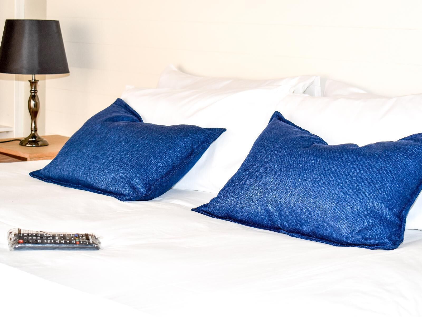 A bed with white linens, two blue pillows, a remote control, and a black lamp on a wooden nightstand.