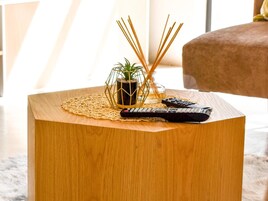 Una mesita auxiliar de madera con un posavasos tejido, una pequeña planta en maceta y dos controles remotos.