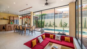 Een ruime woonkamer met een rode zithoek, een eettafel en stoelen. Er is een keuken met kasten en een koelkast. Een groot raam biedt uitzicht op een zwembad en groen.