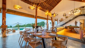 A spacious dining area with a wooden table set for a meal, surrounded by wicker chairs. A staircase with a glass railing leads to an upper level. - Infinity Pool | Private Wellness Space | Sea Views (Lovina)