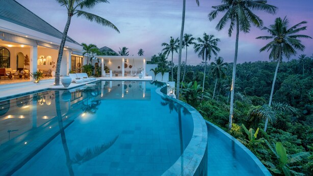 Un área de piscina moderna con sillones de descanso, iluminación interior agradable y vistas a palmeras.