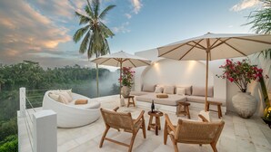 Terraza en la azotea con mobiliario de mimbre, un sofá y sombrillas, con vistas a un paisaje exuberante.