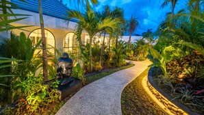 Un sendero conduce a un edificio con ventanas arqueadas, rodeado de exuberante follaje tropical y una estatua decorativa de Buda.
