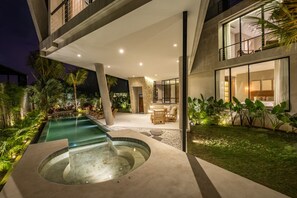 Un área de piscina moderna al aire libre con un jacuzzi, rodeada de exuberante vegetación y un patio bien iluminado con asientos.
