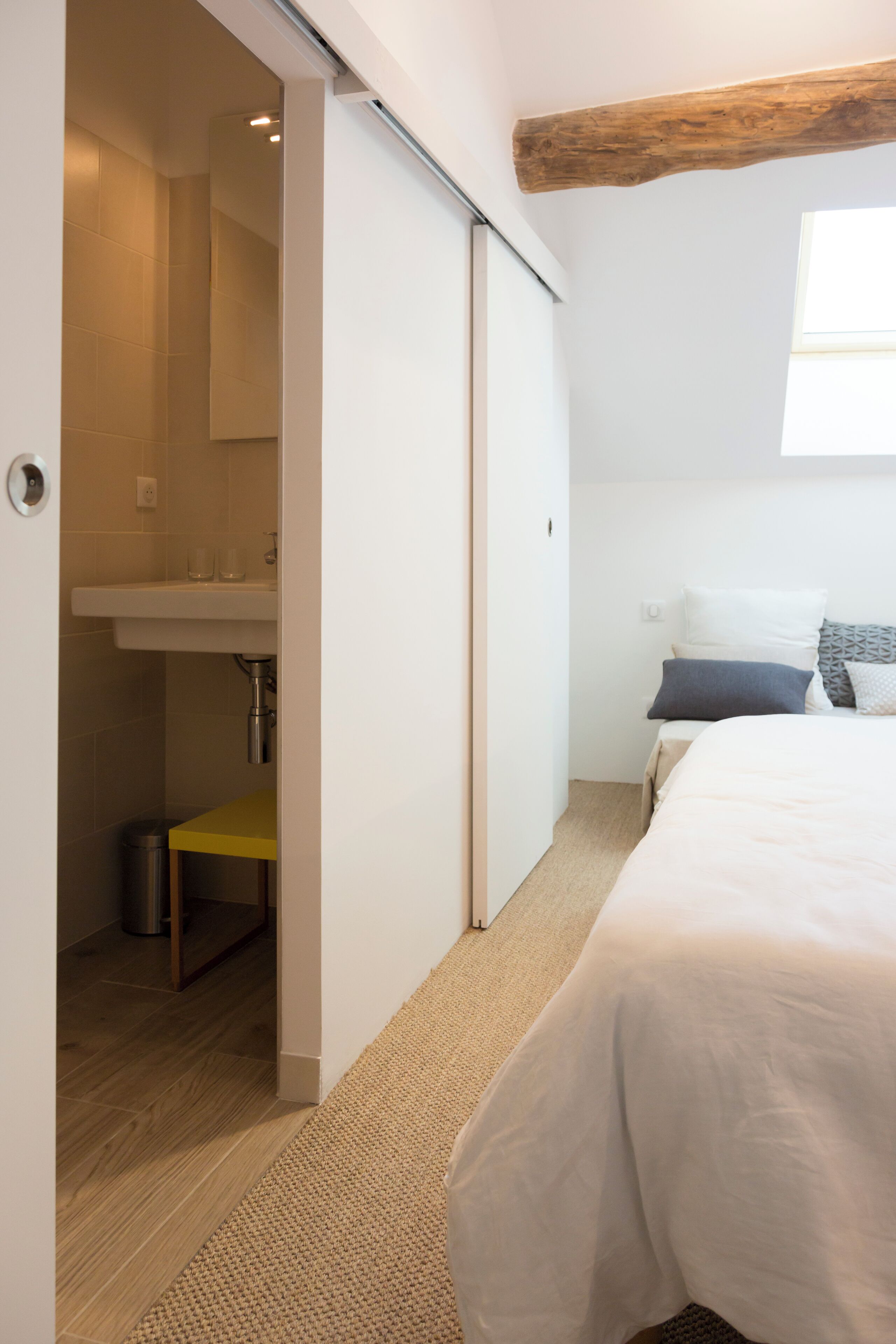 Een slaapkamer met een bed, nachtkastje, raam, houten balk in het plafond, en een badkamer met wastafel en spiegel.
