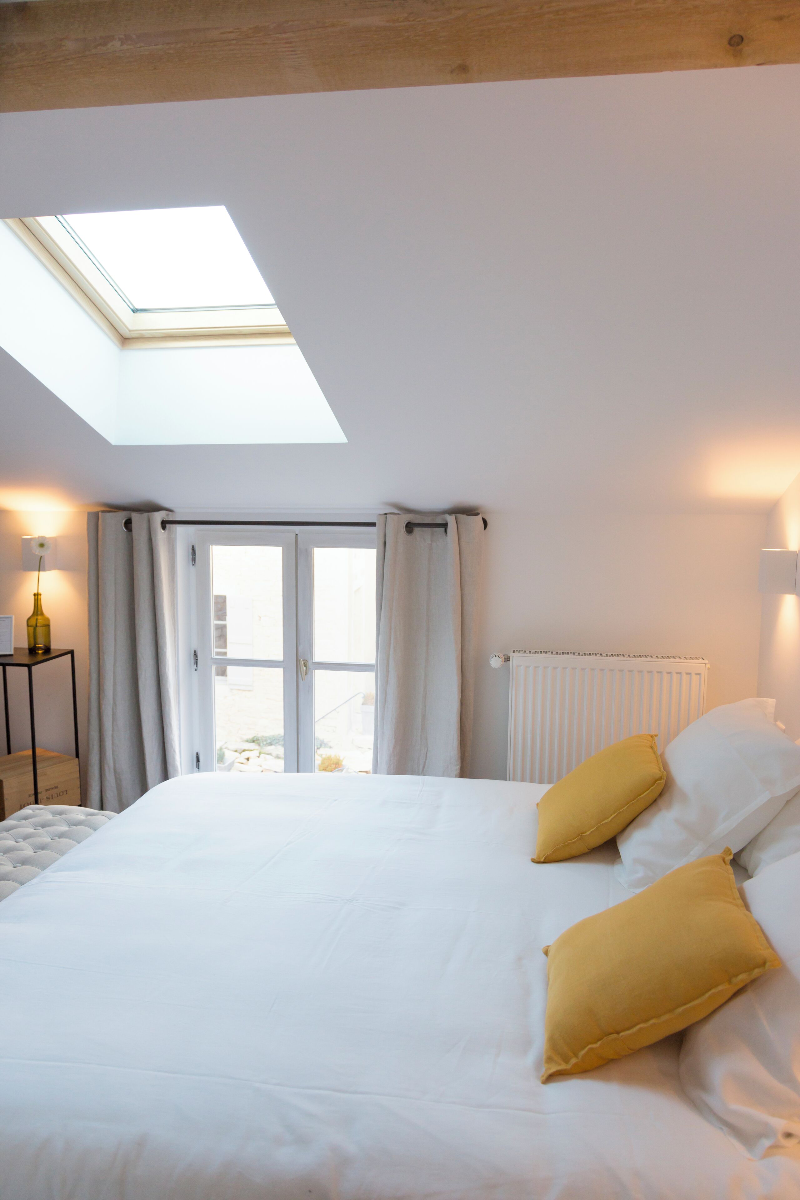 A bedroom with a skylight, a bed with white bedding and two yellow pillows, a bedside table with a lamp, and a radiator.