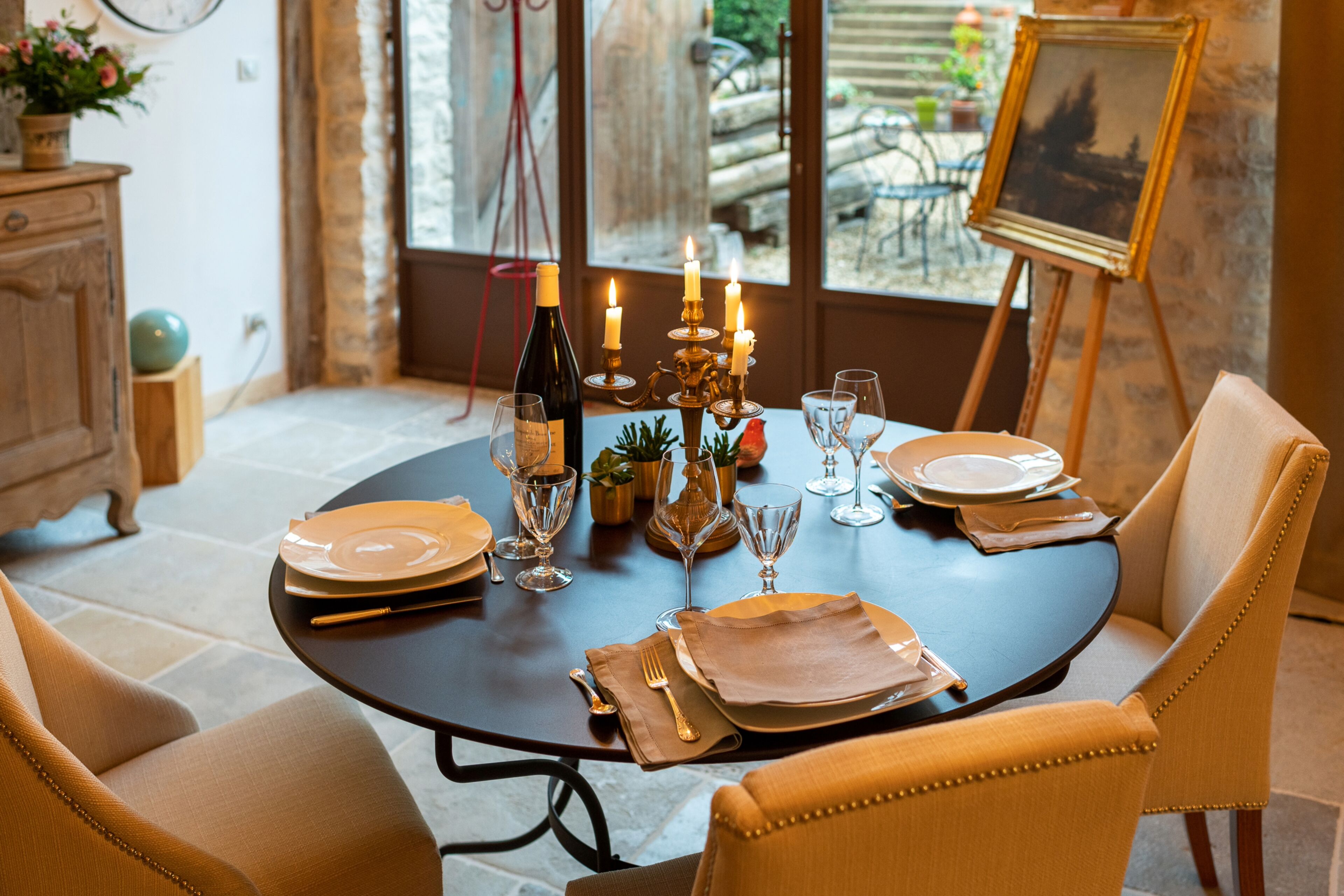 Une table à manger dressée avec des assiettes, des verres et des bougies, entourée de chaises rembourrées.