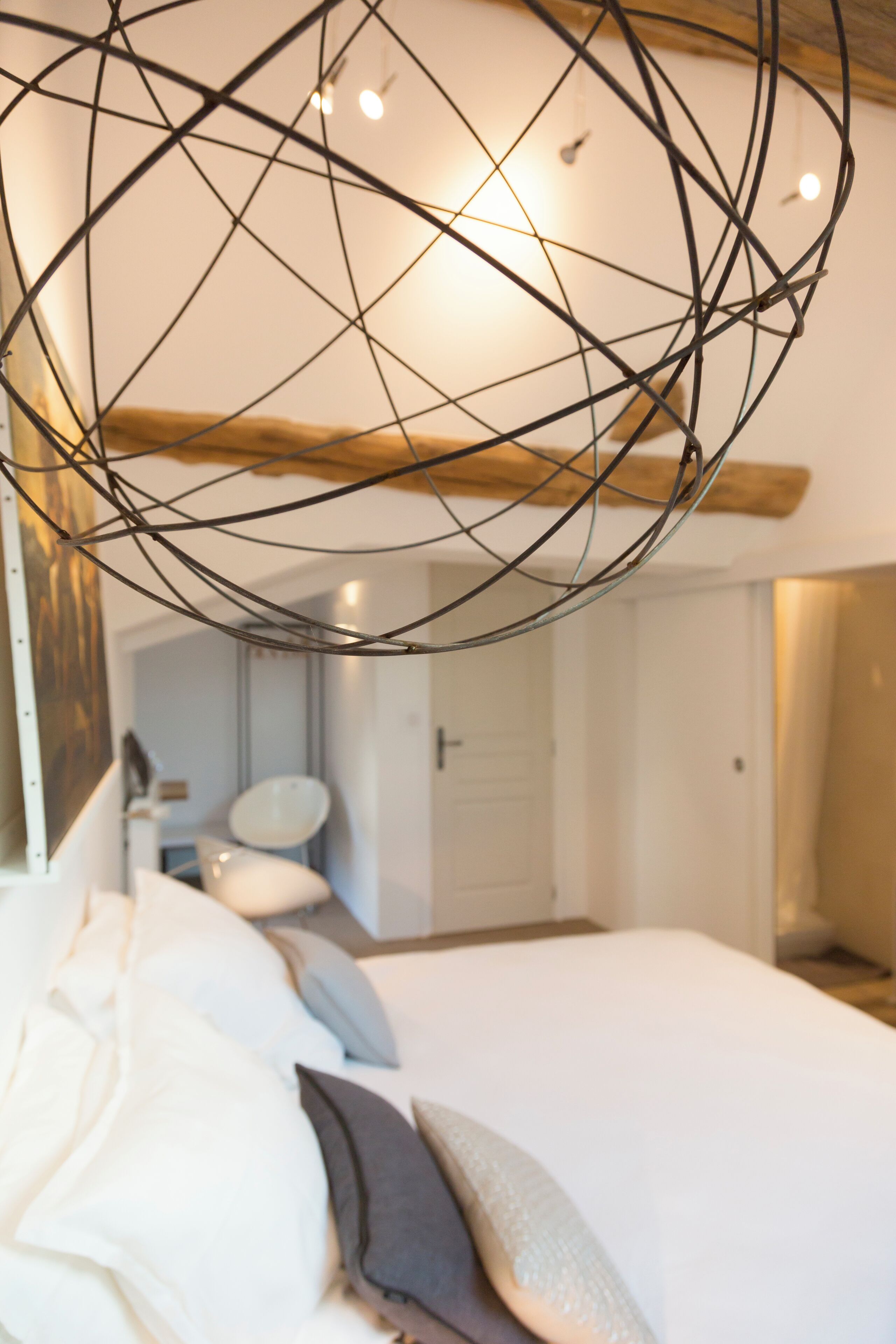 A modern bedroom with a geometric light fixture, a large bed with pillows, and a visible bathroom in the background.