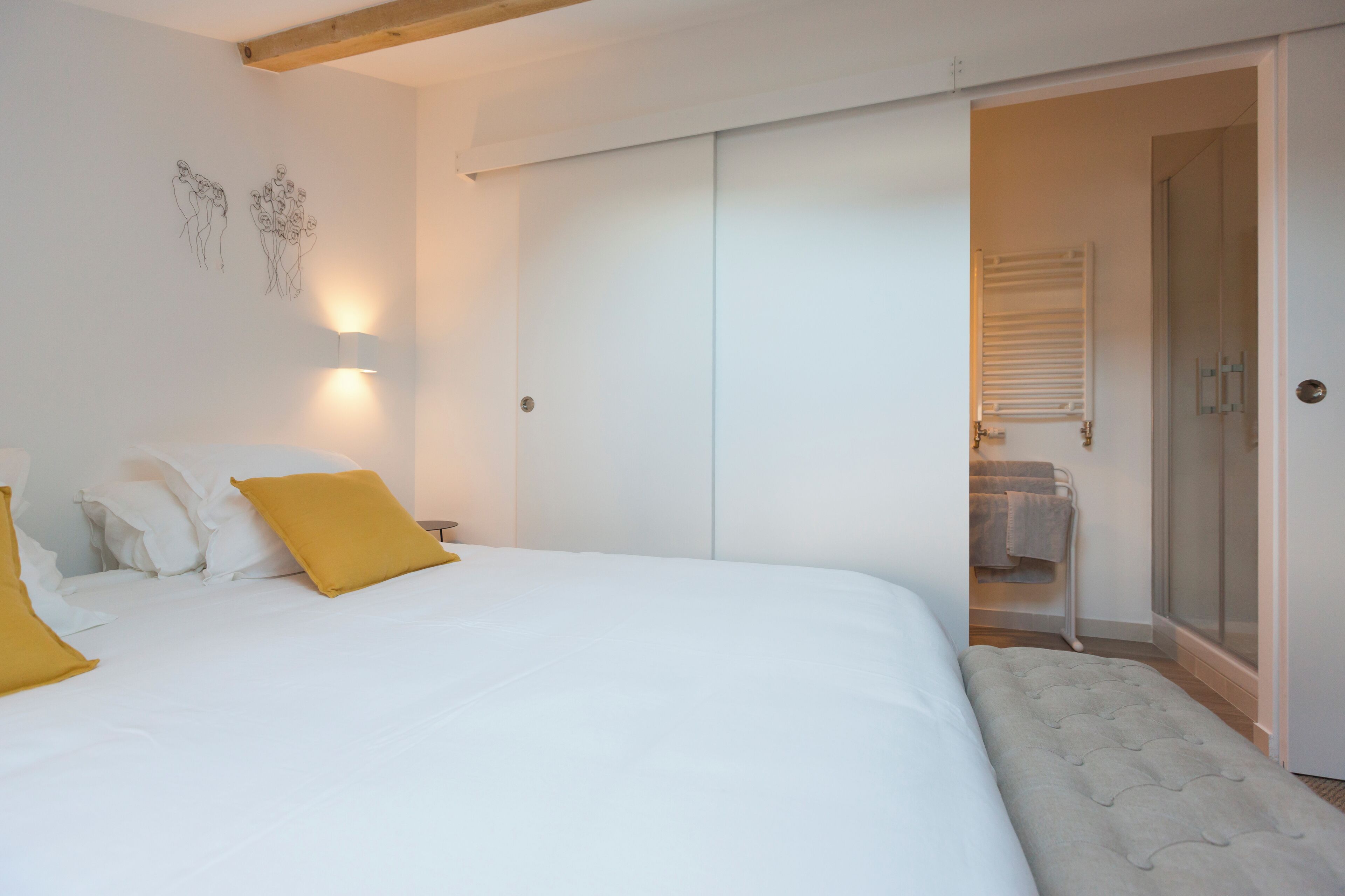 A bedroom with a large bed, a bench, a walk-in closet, and a bathroom with a shower and towel rack.