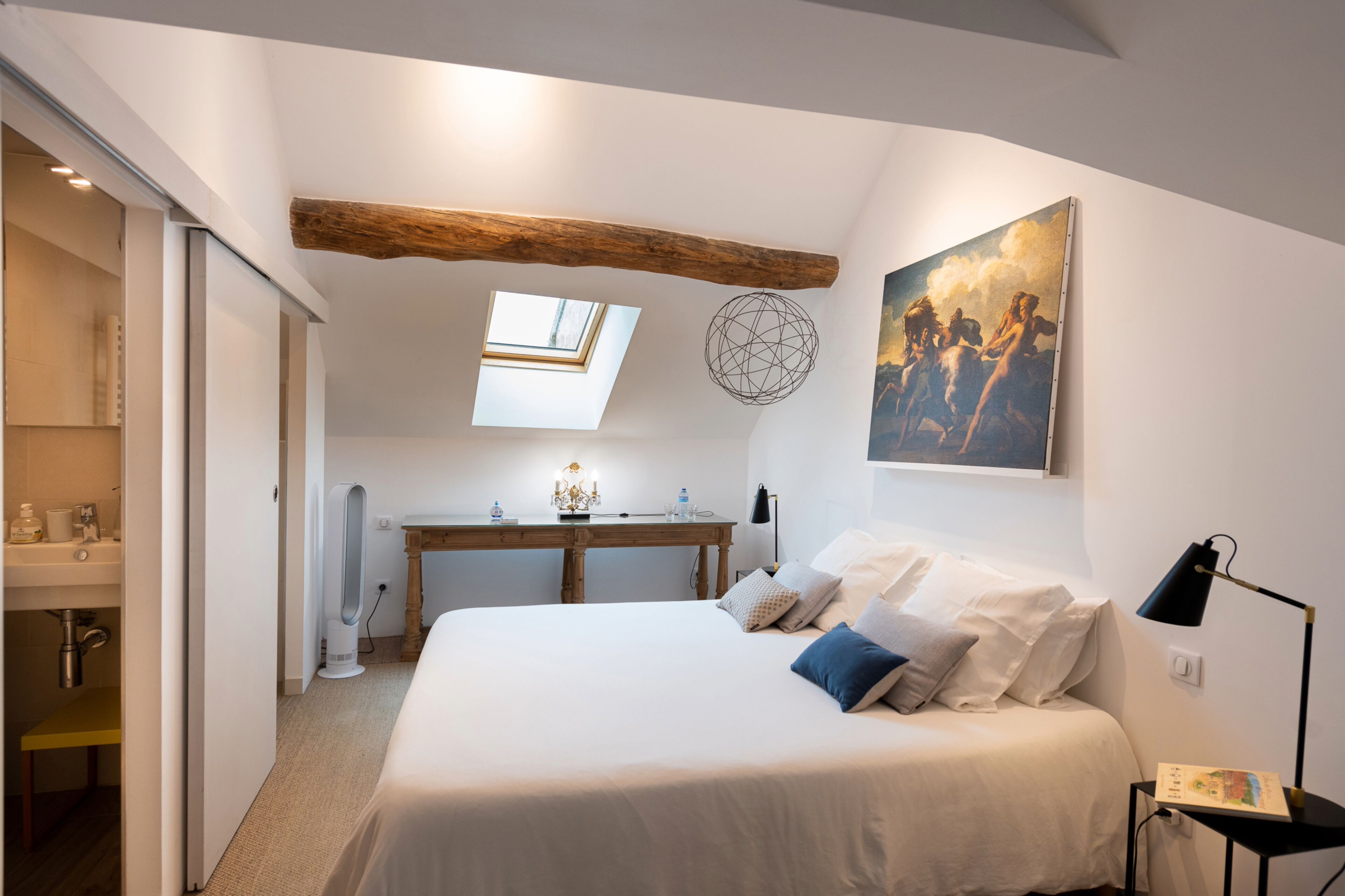 Une chambre avec un grand lit, un bureau, une lucarne et un tableau au mur.