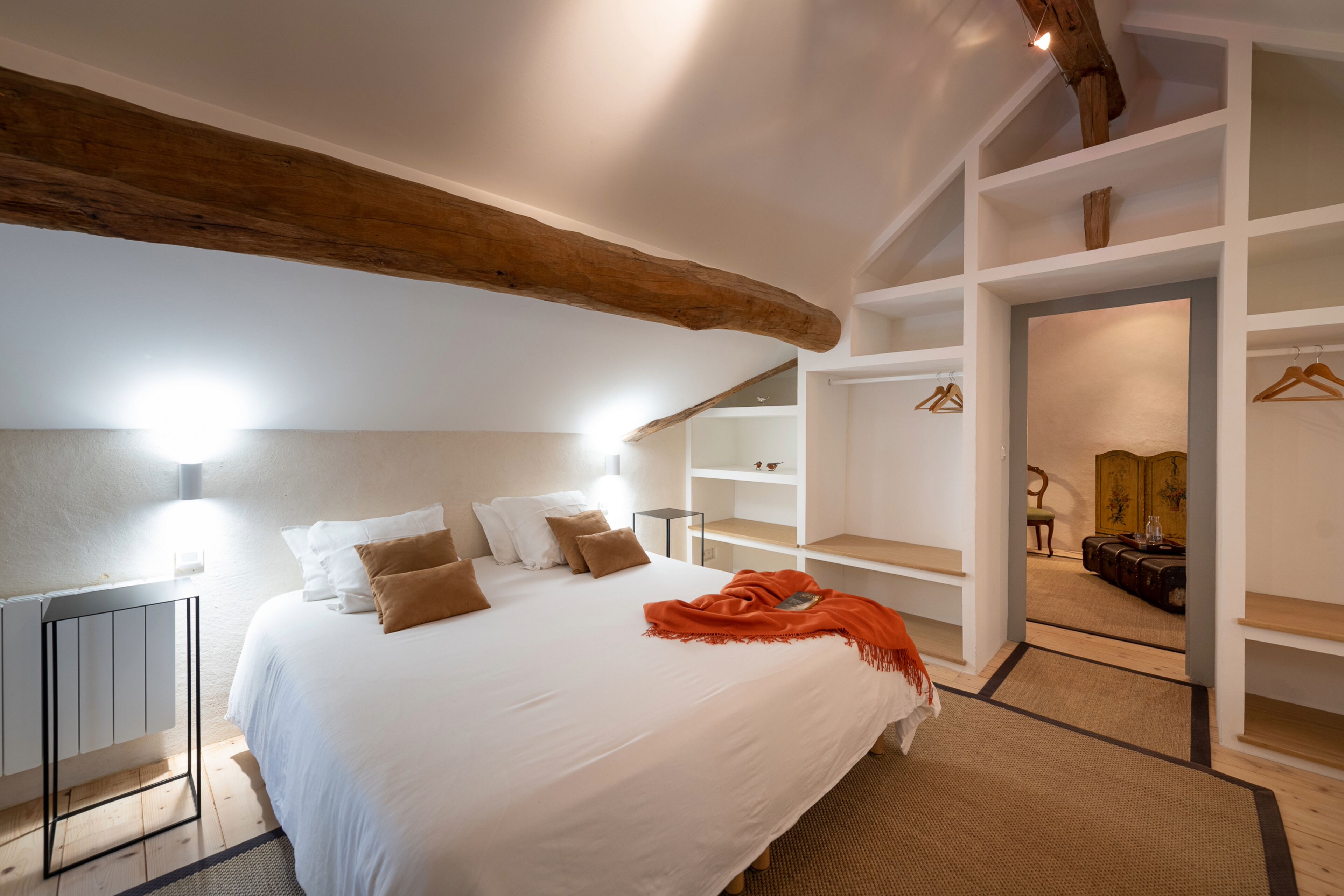 Une chambre avec un grand lit, des murs blancs et des poutres en bois. On y trouve un dressing avec des étagères et une chaise.