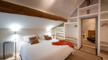 Une chambre avec un grand lit, des murs blancs et des poutres en bois. On y trouve un dressing avec des étagères et une chaise.