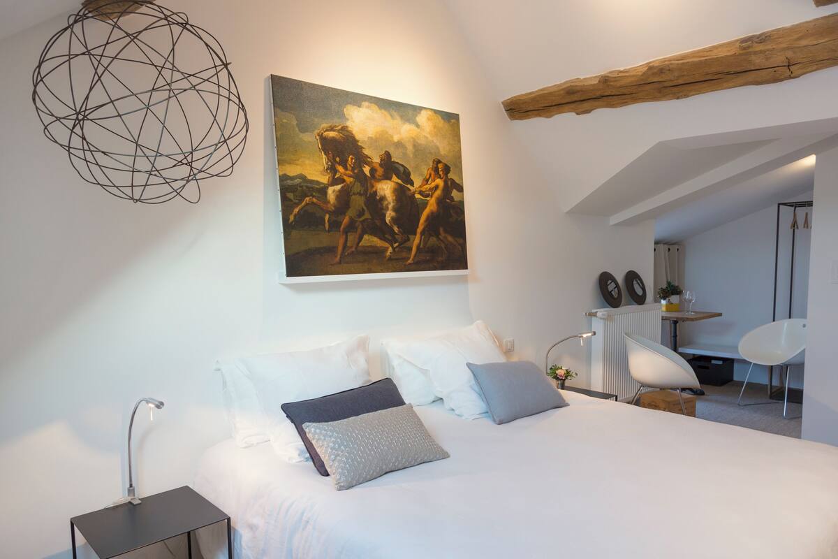 A modern bedroom with a large bed, a desk, and a chair. A painting is on the wall, and there is a unique spherical light fixture.